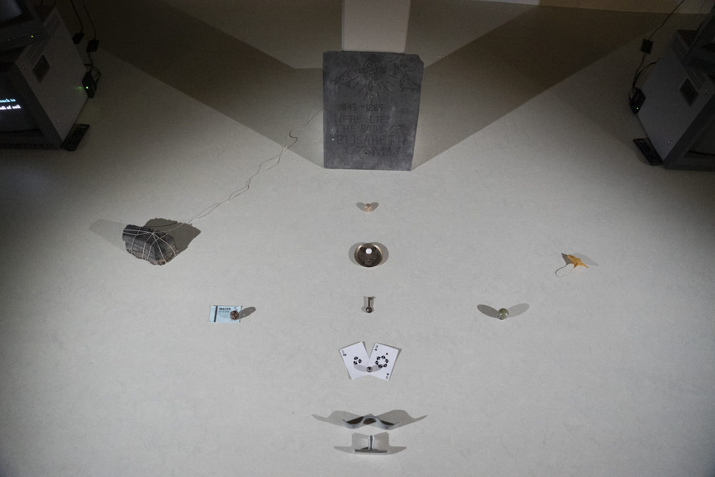 This image looks at objects laid out in a geometric pattern on the floor, such as playing cards, a rock tied with string, a small key, a leaf in front of a mock gravestone